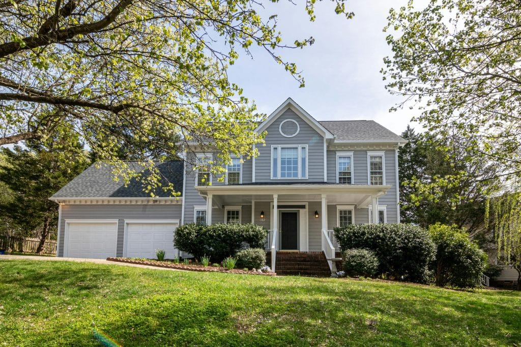 Two-story gray house with white trim, a front porch, and a three-car garage, surrounded by trees and a sloped green lawn.