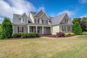 A large two-story suburban house with light-colored vinyl siding, red shutters, and a gray shingle roof, surrounded by neatly trimmed shrubs, mulch beds, and a well-maintained lawn under a partly cloudy blue sky.