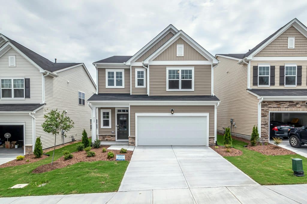 A modern two-story suburban house with tan vinyl siding, white trim, and a double garage door, flanked by similar neighboring homes. The front yard features a freshly landscaped lawn with small shrubs, mulch beds, and a young tree, set against a cloudy sky.