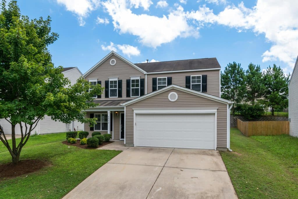 Two-story suburban house with beige siding, black shutters, white garage door, and a clean driveway, surrounded by green grass, trimmed shrubs, and a tree under a partly cloudy blue sky.