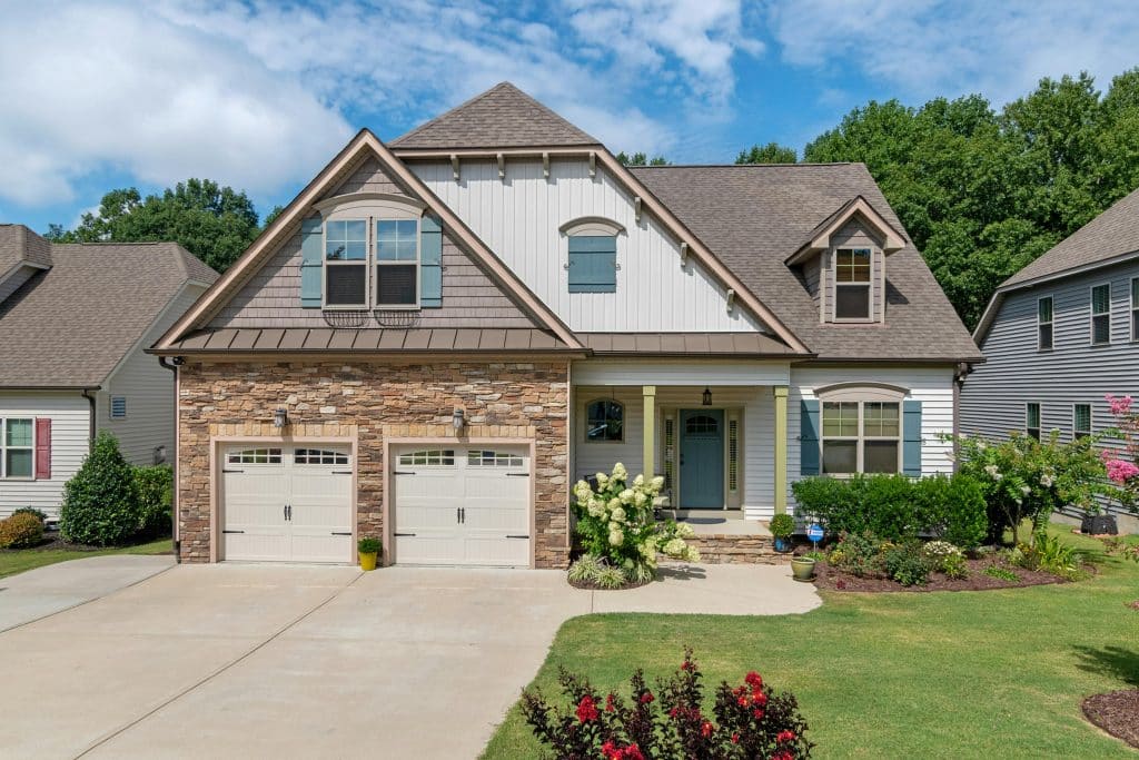 Modern suburban home with stone and siding exterior, two-car garage, manicured lawn, blooming flowers, and decorative shutters under a partly cloudy sky.