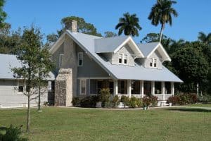 A well-maintained gray house with a stone chimney, white trim, and a spacious front porch, surrounded by palm trees and a manicured lawn under a clear blue sky.