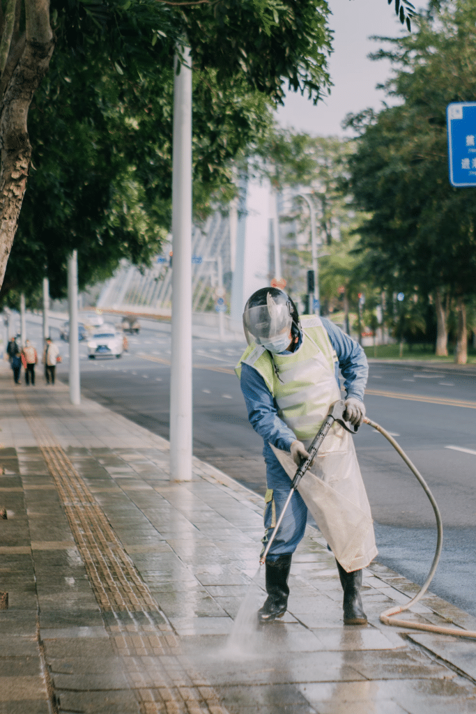 Worker in protective gear pressure washing a city sidewalk, removing dirt and grime from the pavement near a tree-lined urban street.
