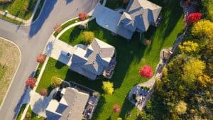 Aerial view of suburban homes with well maintained lawns, driveways, trees with fall colors, and a wooded area bordering the neighborhood.