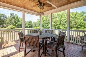 A covered outdoor deck with wooden flooring, a round wooden table, and several matching chairs, surrounded by white railings and overlooking a green landscaped yard.